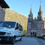 Vista fronto lateral de una furgoneta preparada para llevar pasajeros, donde se ve el logo «Taxitur», sobre el texto «Lagos de Covadonga», tanto en la parte frontal como en el lateral de la misma. Al fondo vemos la Basílica de Covadonga.