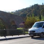 Vista de una furgoneta preparada para llevar pasajeros, donde se ve el logo «Taxitur», sobre el texto «Lagos de Covadonga», tanto en la parte frontal como en el lateral de la misma. Al fondo vemos el puente de Cangas de Onis, con la réplica de la Cruz de la Victoria colgando de el