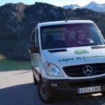 Vista frontal de una furgoneta preparada para llevar pasajeros, donde se ve el logo «Taxitur», sobre el texto «Lagos de Covadonga». Al fondo vemos una panorámica de los Lagos de Covadonga