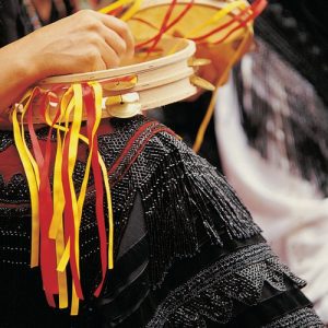 Primer plano de la falda de un traje regional de Llanes, sobre la rodilla se puede ver una pandereta con lazos rojos y amarillos