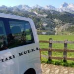 Vista parcial de la parte trasera de una furgoneta preparada para llevar pasajeros, donde se ve el texto «Taxi Huera»: Al fondo una panorámica de los Picos de Europa