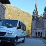 Vista parcial de una furgoneta preparada para llevar pasajeros, donde se ve el logo «Taxitur», sobre el texto «Lagos de Covadonga», tanto en la parte frontal como en el lateral de la misma. Al fondo vemos la Basílica de Covadonga