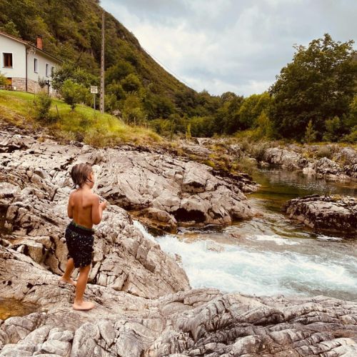 Un niño observa una poza natural en el río Dobra, sobre unas rocas que rodean la poza. A un lado vemos una casa pintada de blanco y al fondo una arboleda y la ladera verde de un monte